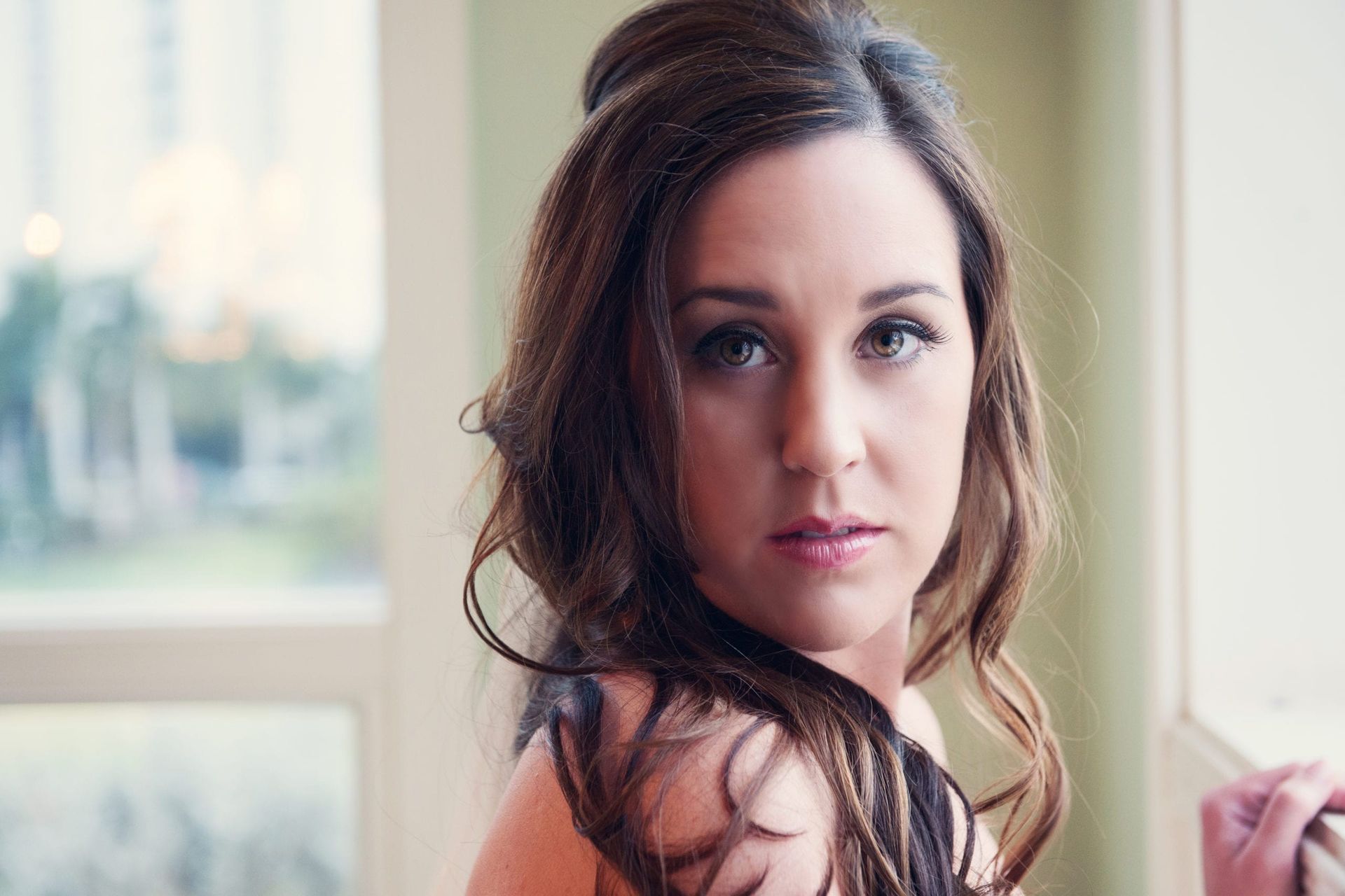 Woman with long brown hair, looking towards the camera, soft focus with window backdrop.