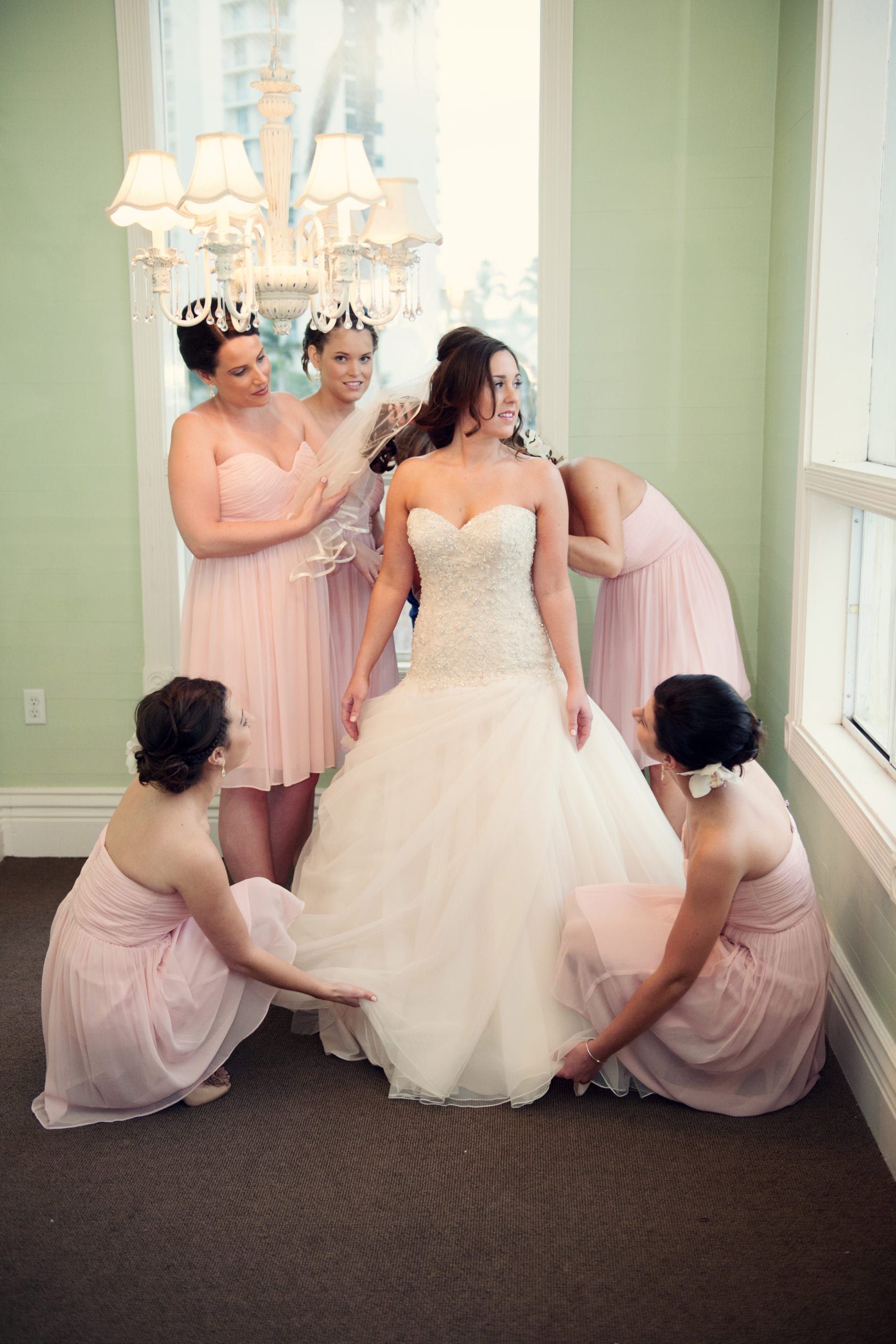 Bride surrounded by bridesmaids in pink dresses, adjusting the train of her white wedding gown.