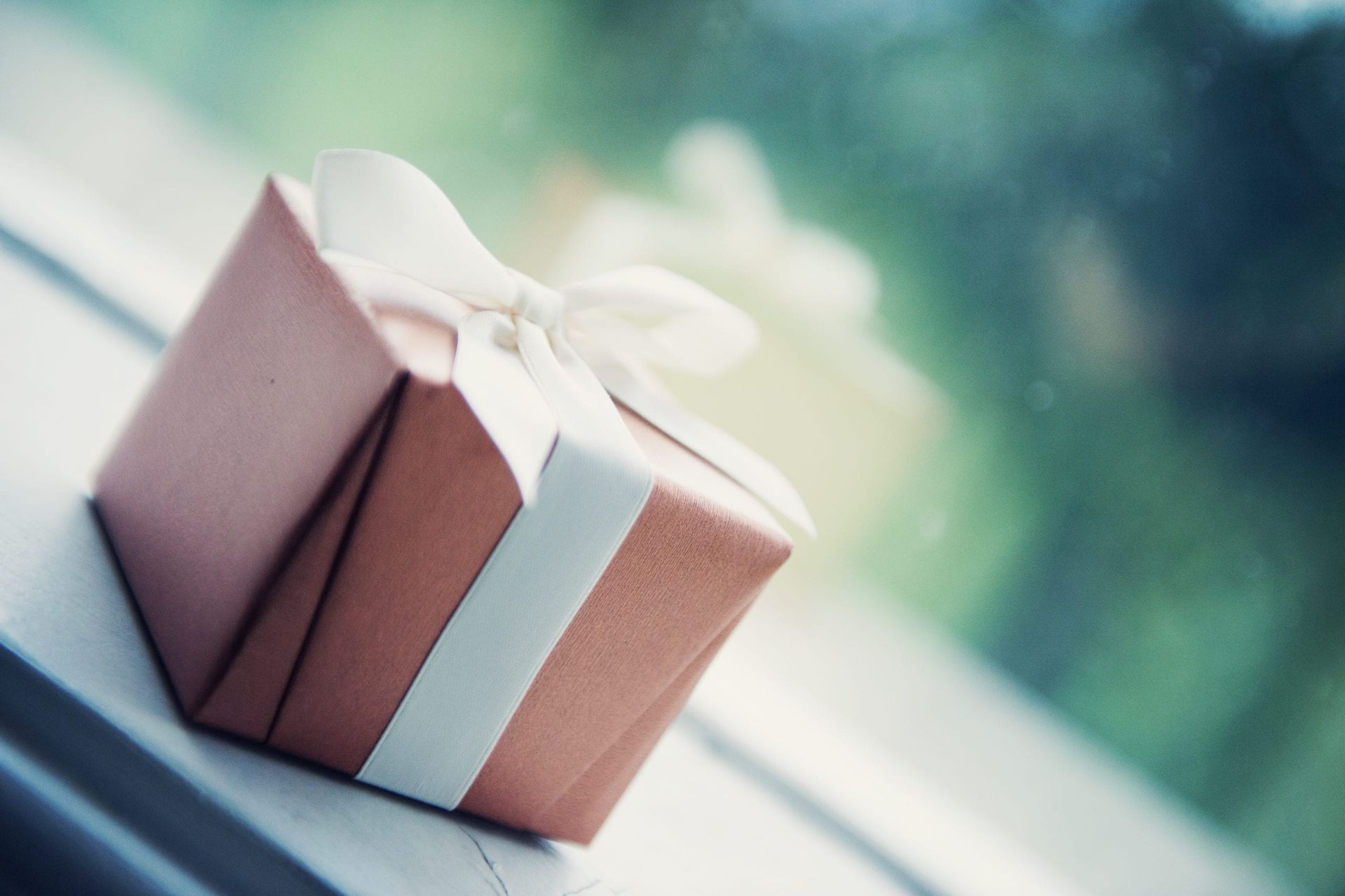 Brown gift box with white ribbon on a windowsill, green blurred background.