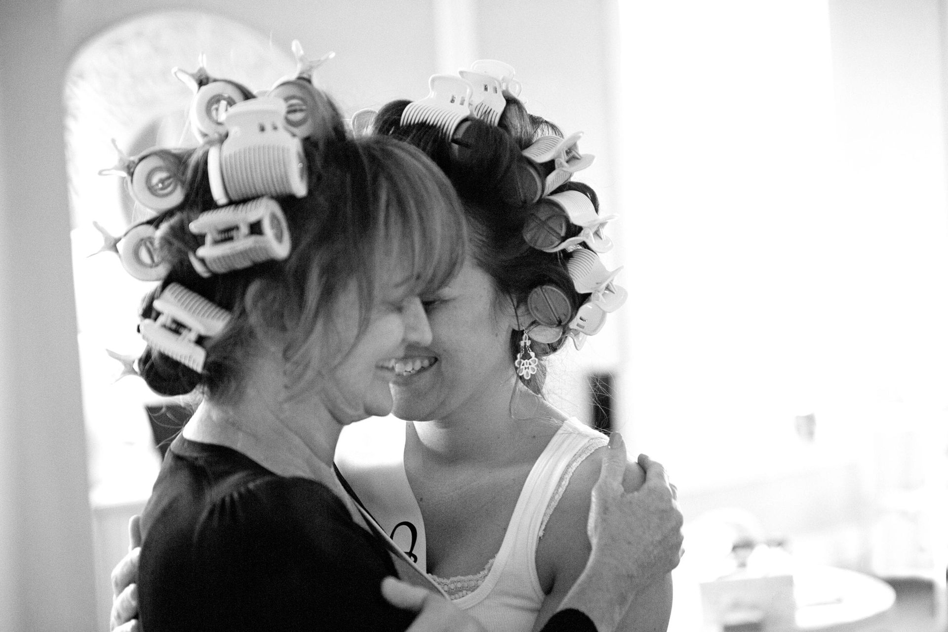 Two women embracing, both with curlers in their hair, smiling.