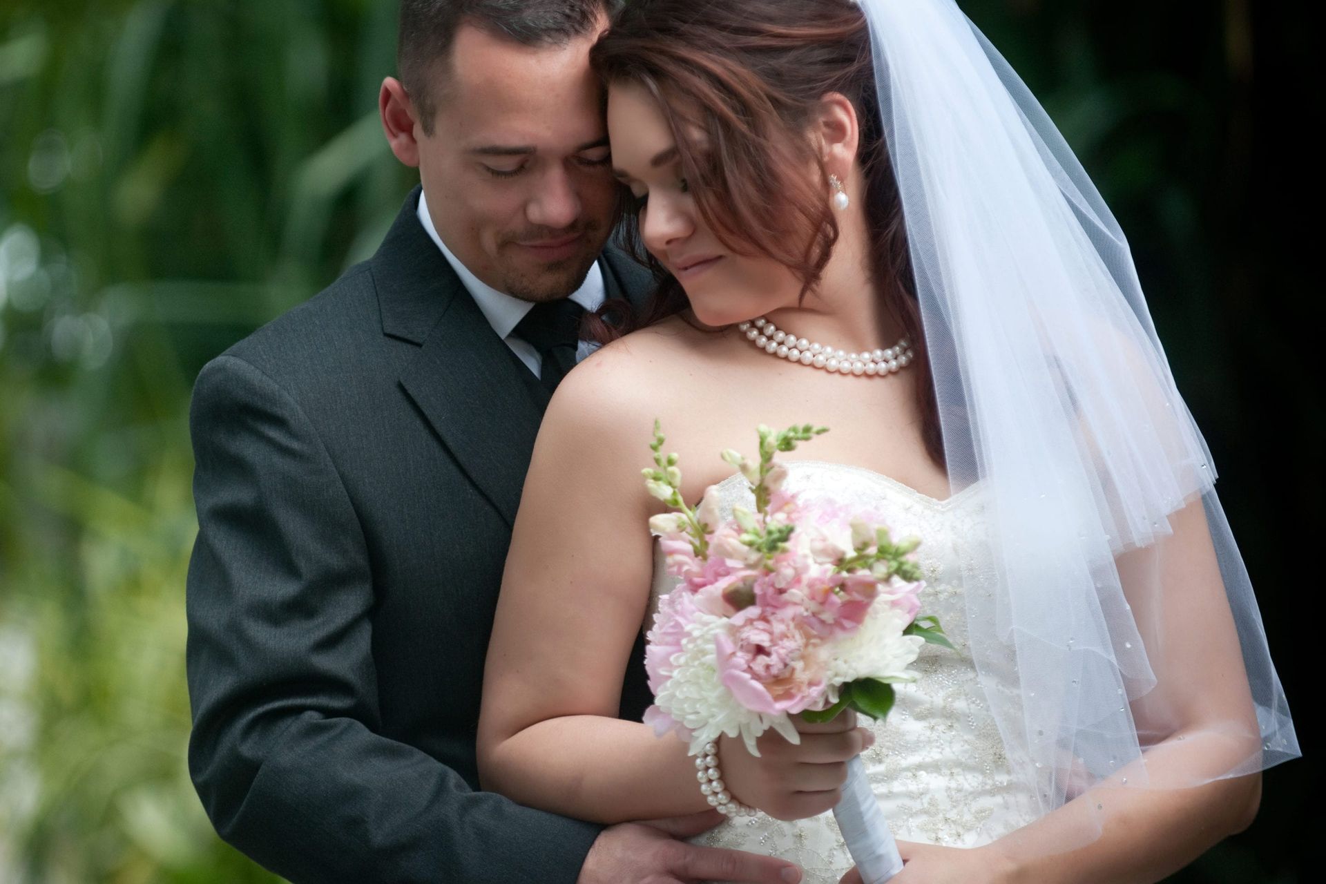 Bride and groom embracing, she in white dress and veil, holding flowers; he in suit. Outdoor setting, soft focus.