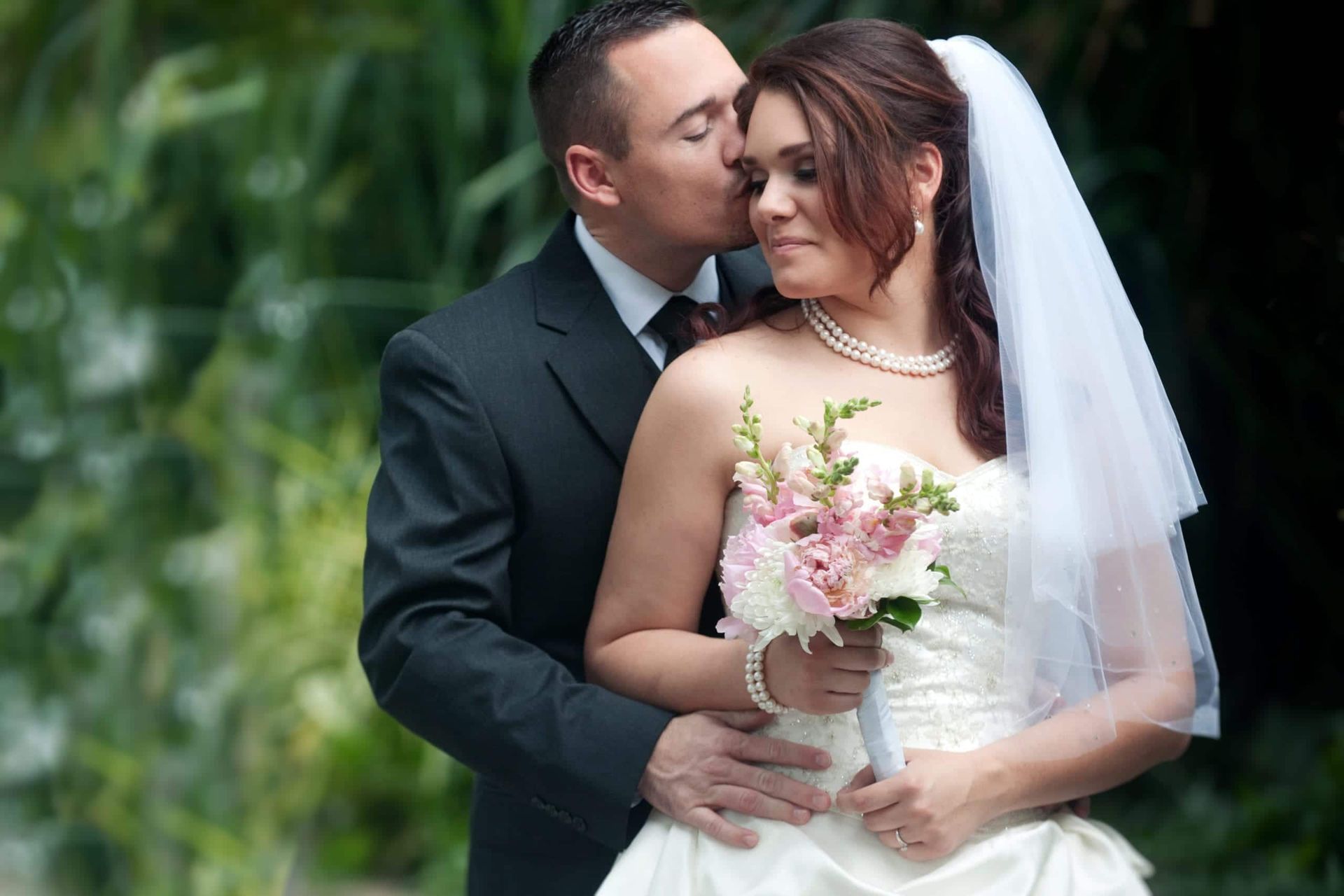Groom kissing bride's cheek. Bride holds flowers, wearing a white dress and veil. Lush green background.