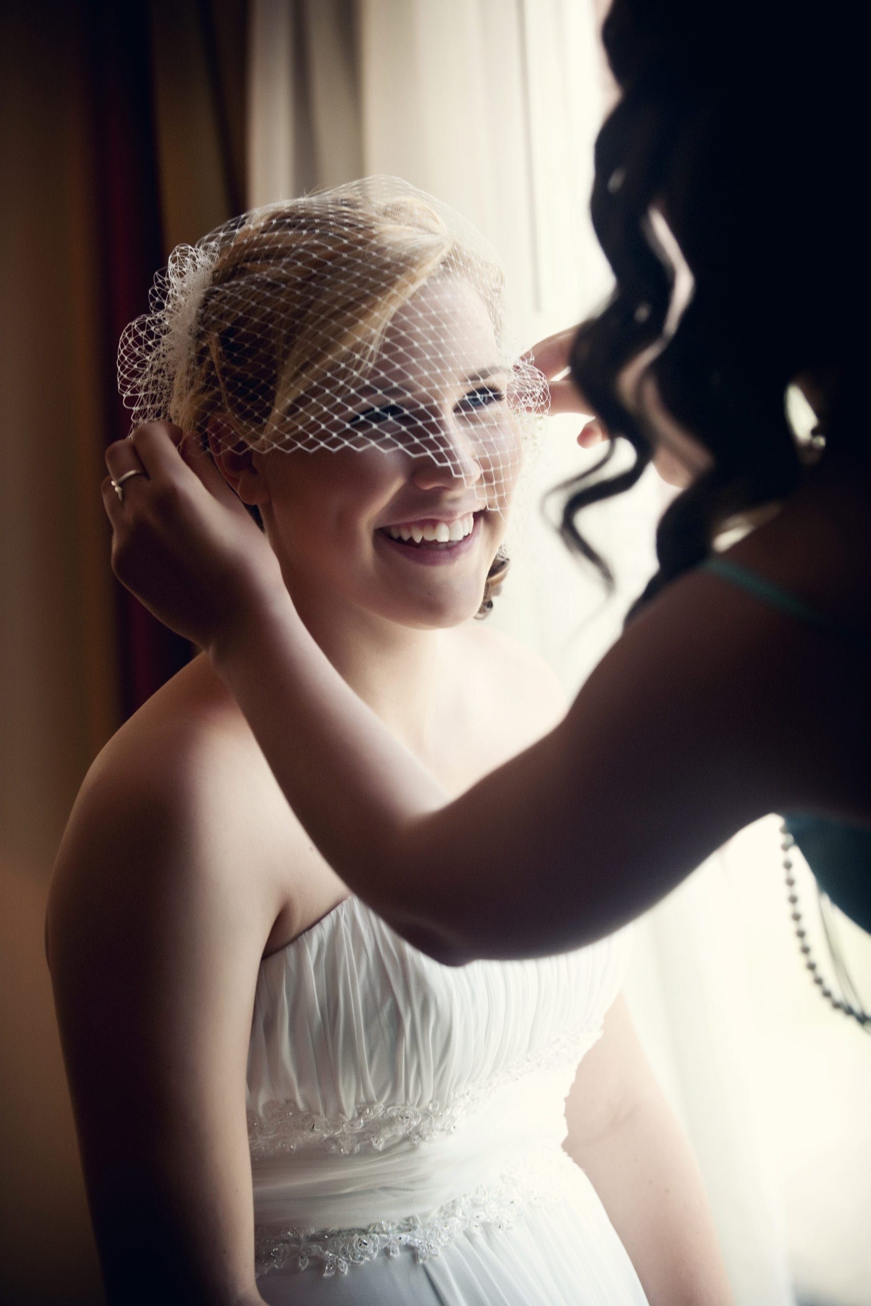 Bride smiling as bridesmaid adjusts her veil. Sunlight streams in window.