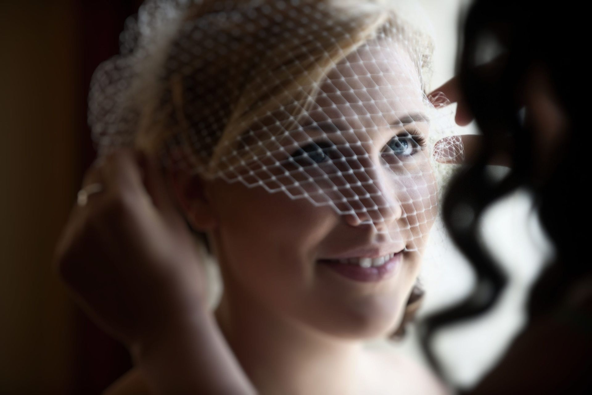 Bride smiling, wearing a veil, as someone adjusts it. Indoors, natural light, focus on her face.