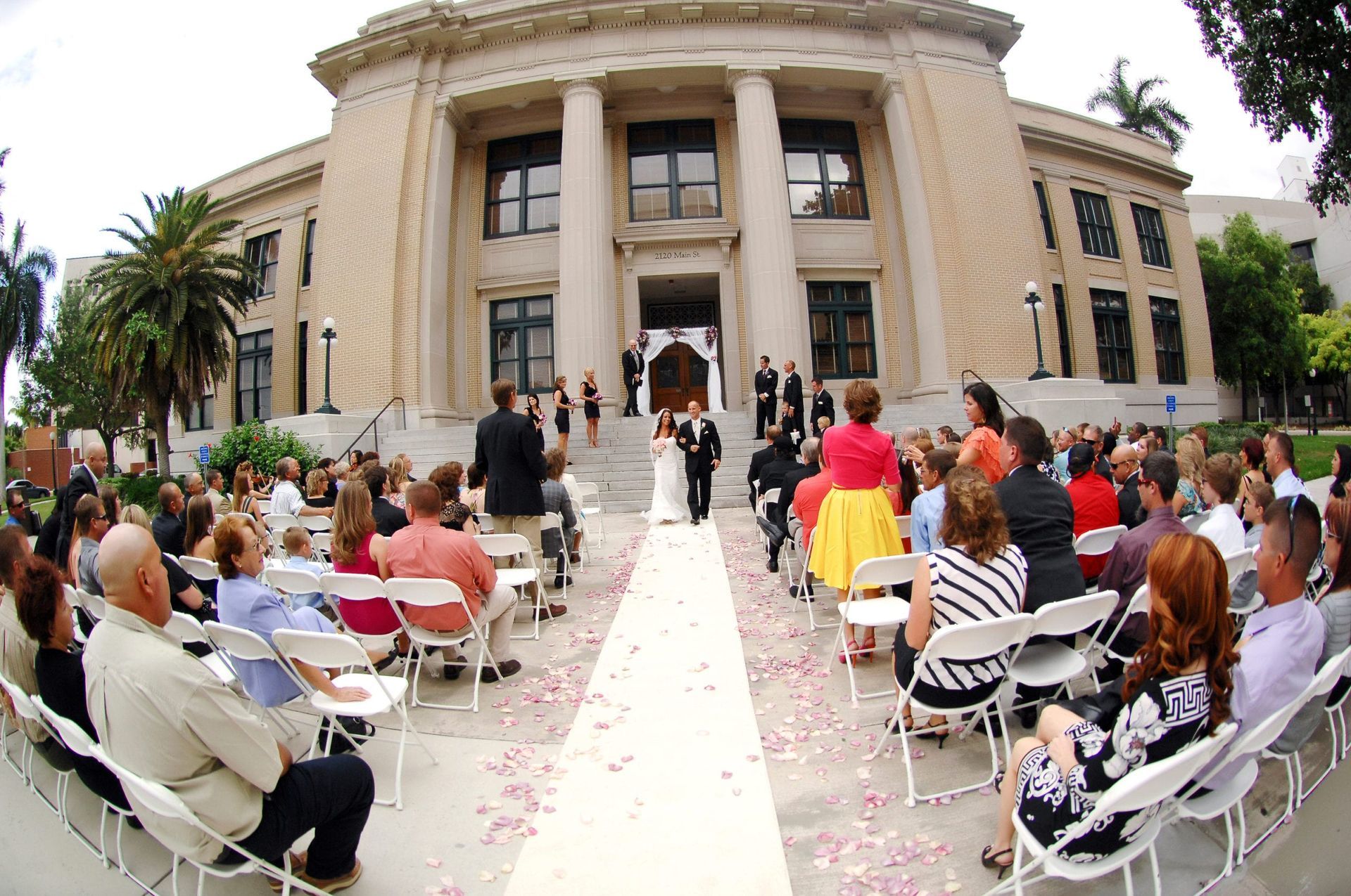 Wedding ceremony outside a large, historic building. Bride and groom walk down aisle lined with guests.