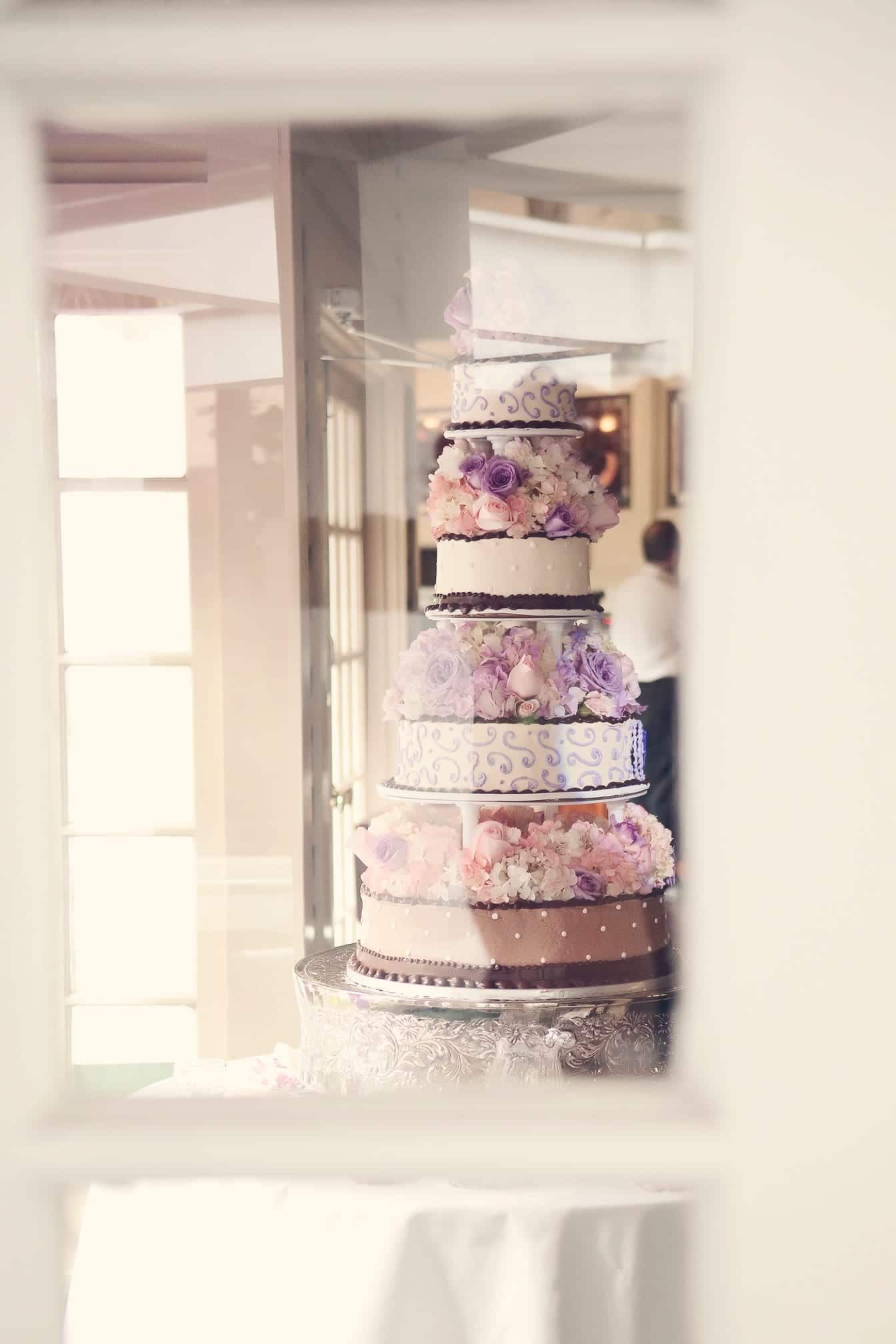 Six-tier wedding cake with pastel floral accents, viewed through a window.