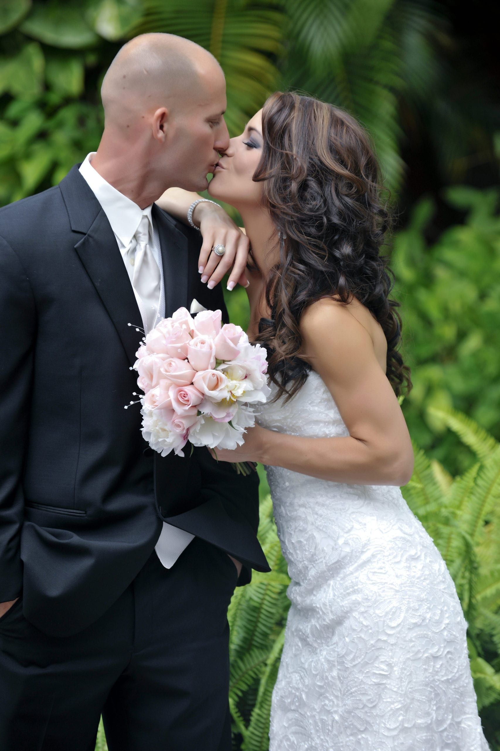 Bride and groom kissing outdoors, she in white dress, he in black suit, holding pink bouquet.