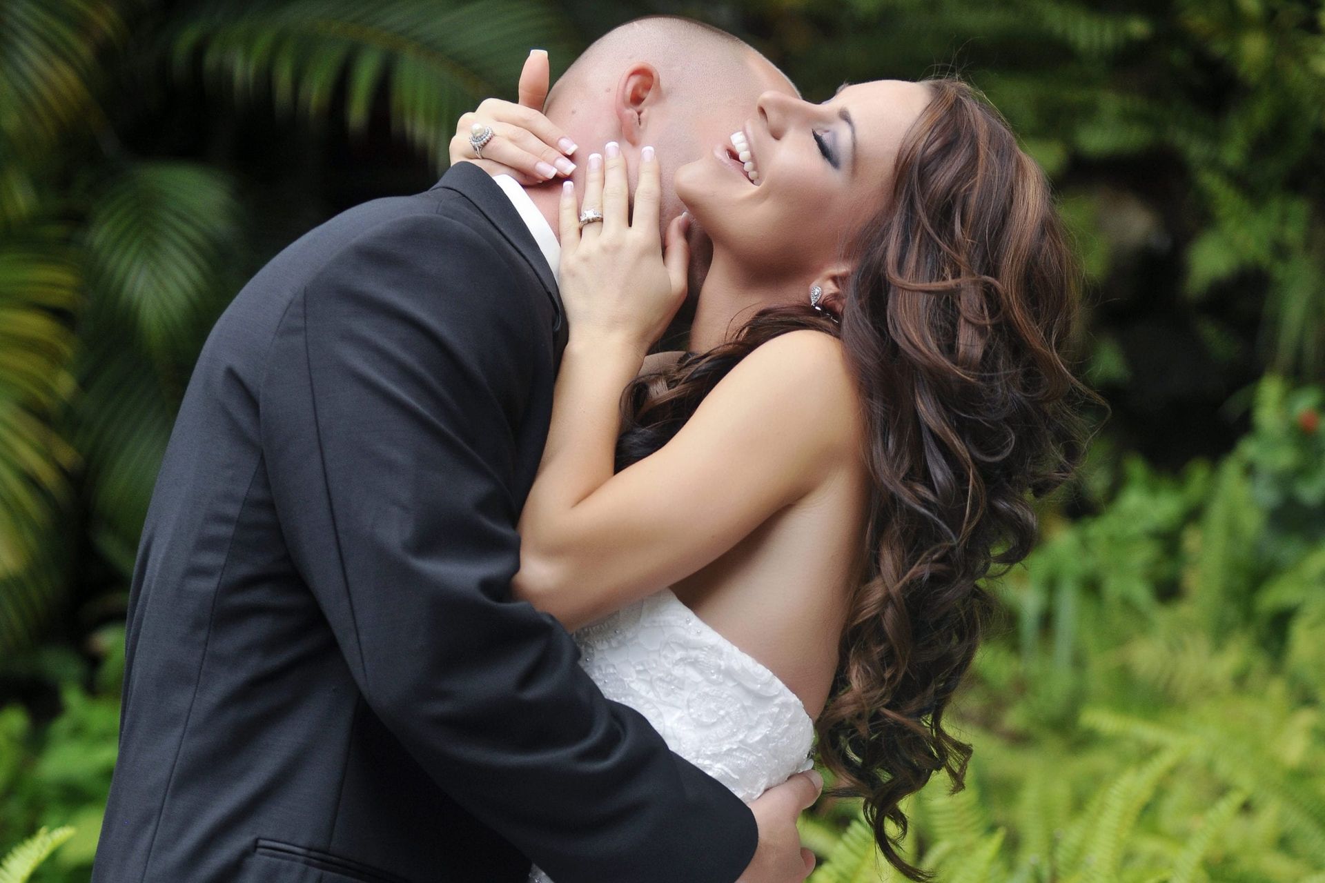 Bride and groom embrace and laugh outdoors; he kisses her neck.