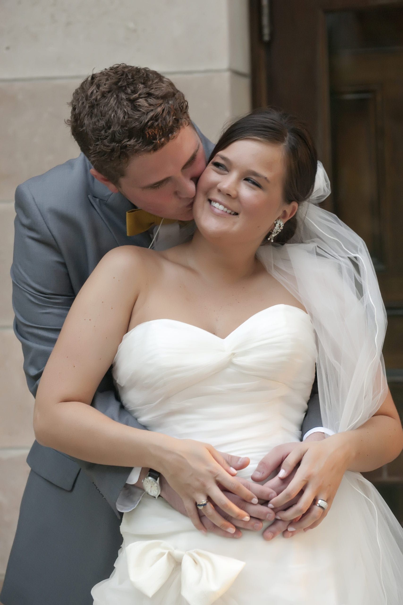 Groom kissing bride's cheek; she smiles, wearing a white strapless dress, with his arm around her waist, outside a building.