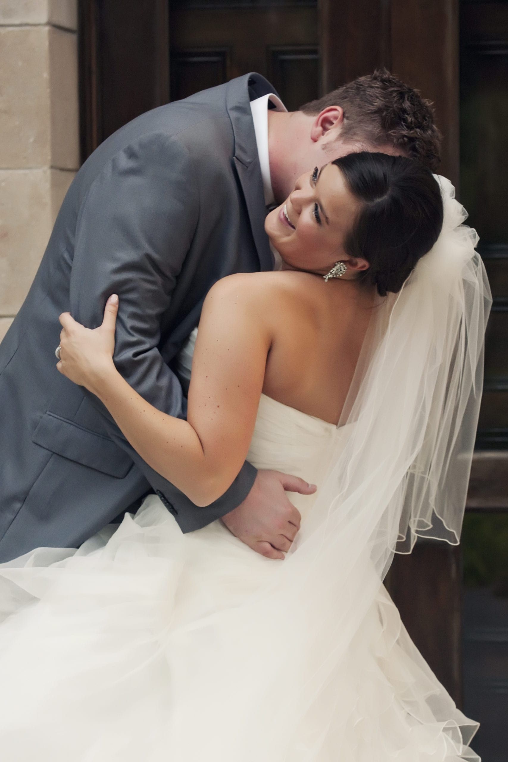 Bride and groom embrace, groom kisses bride's neck. He wears gray suit. She wears a white strapless dress, veil.