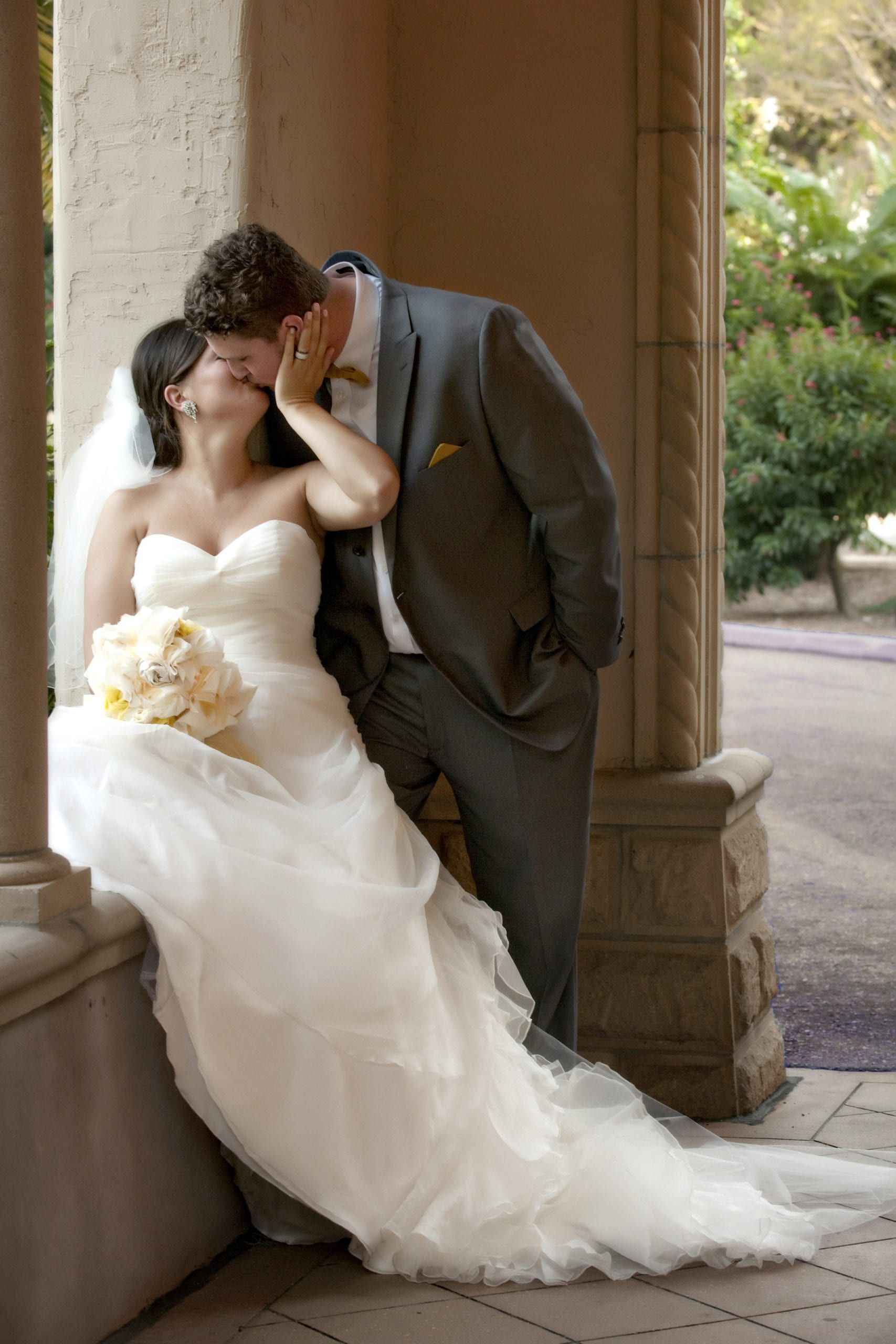 Bride and groom kissing, bride wearing white strapless dress, groom in grey suit, in a stone archway.