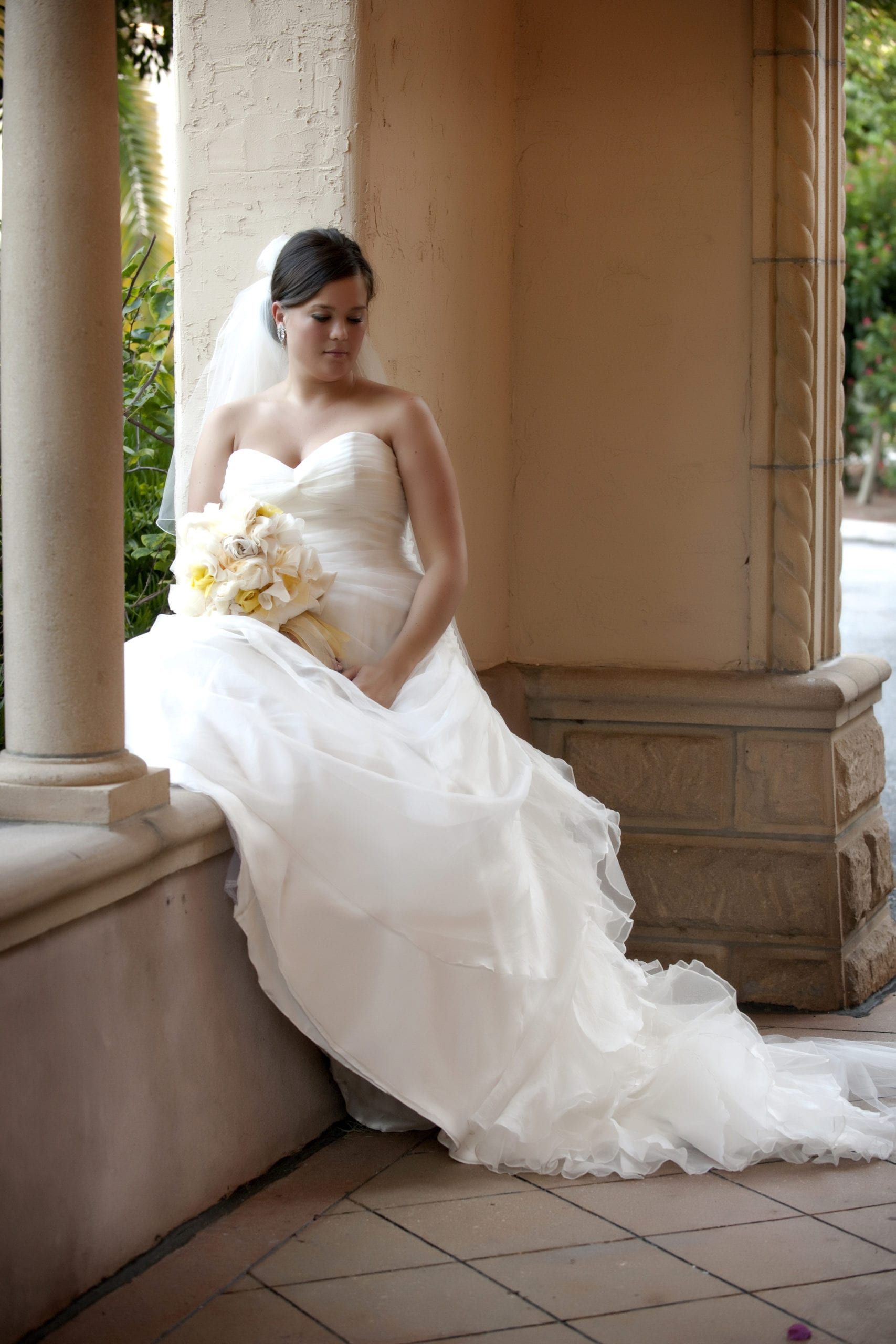 Bride in strapless white dress sits on a stone ledge, holding a bouquet. Light, airy setting.