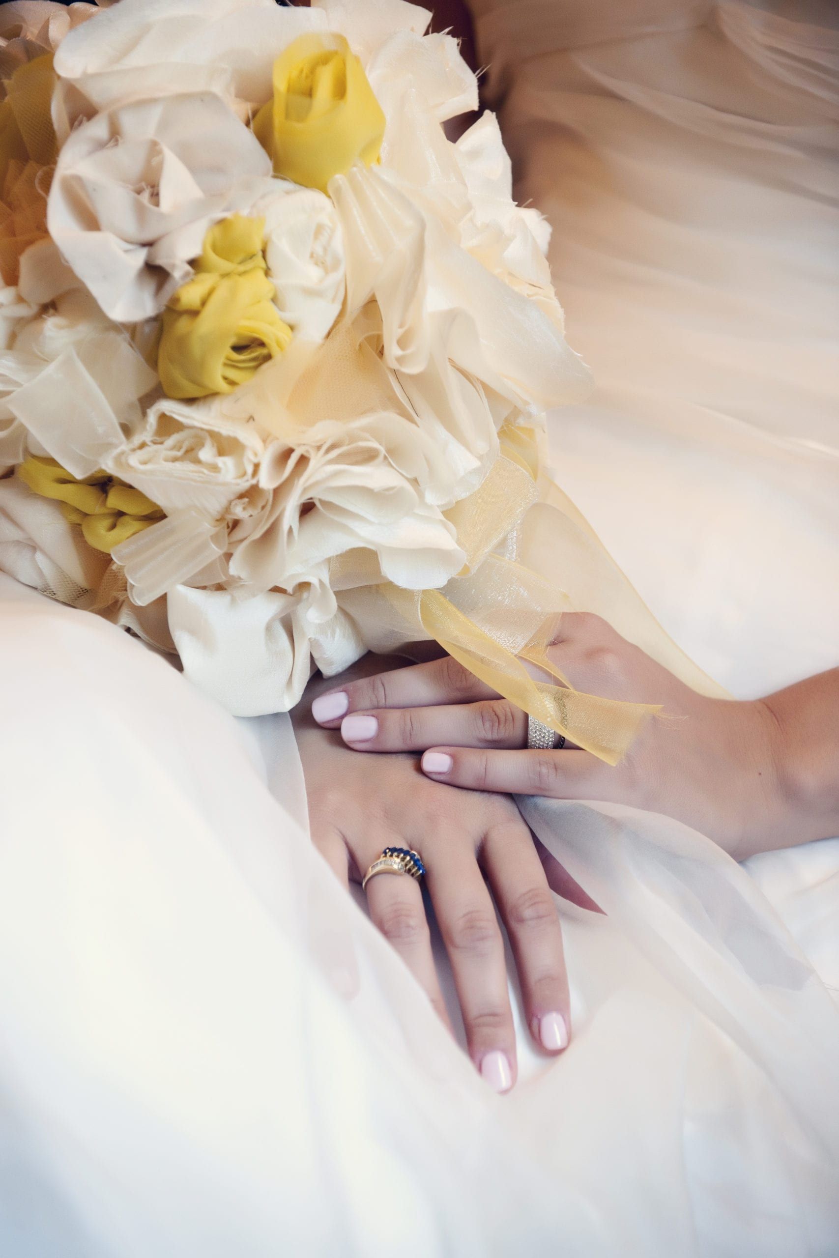 Bride's hands with rings, holding a bouquet of yellow and cream-colored flowers, resting on her dress.