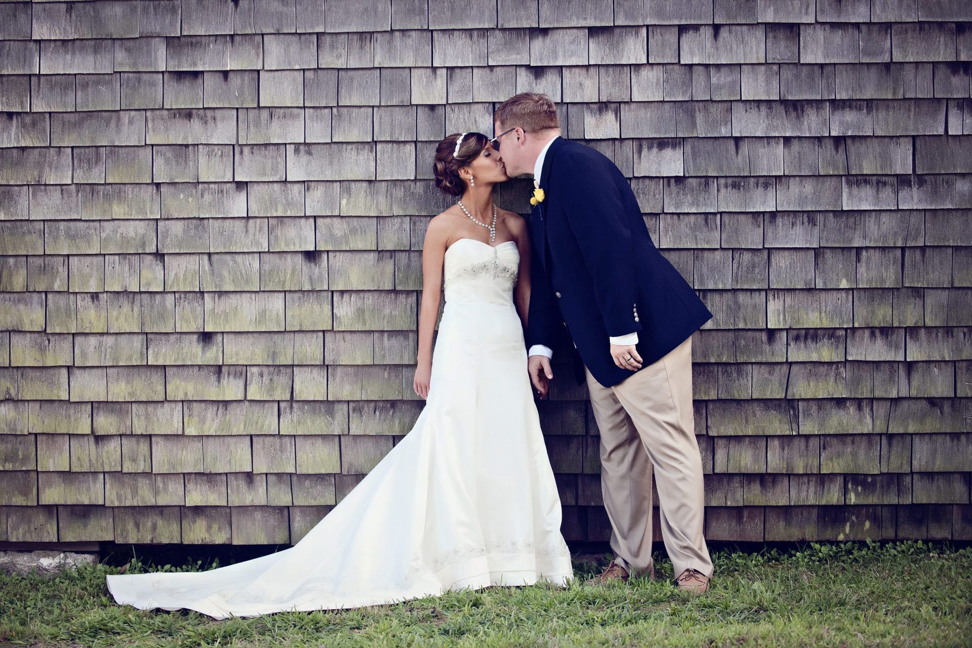 Bride and groom kissing in front of a weathered wooden wall. The bride wears a strapless white gown, the groom a blazer and khaki pants.