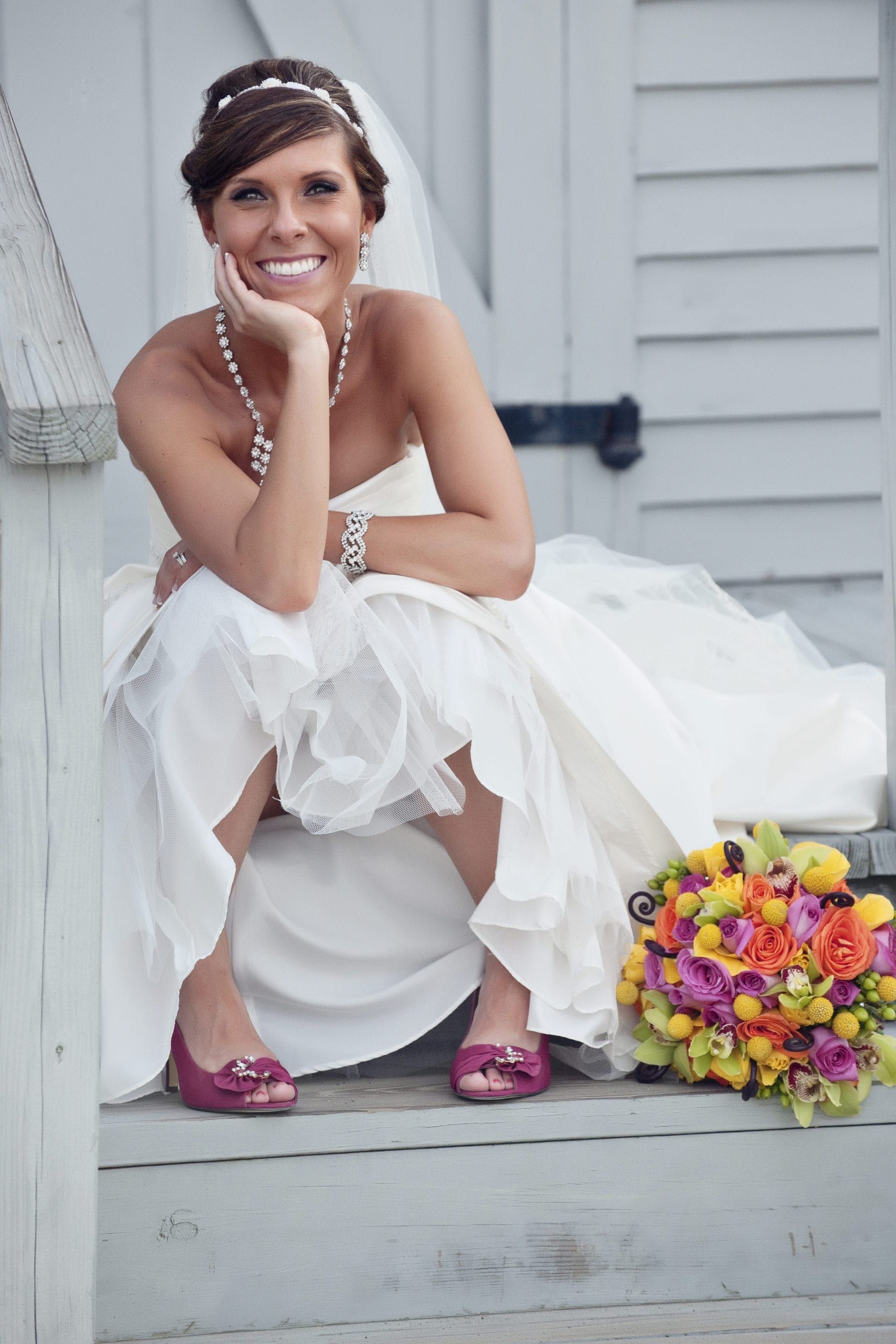 Bride in white dress sits on steps, smiling, with bouquet and pink shoes.