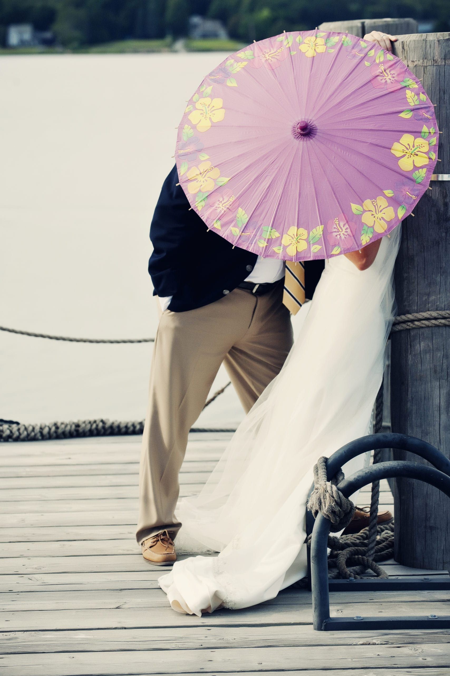 Bride and groom hide under a purple parasol on a wooden dock, near water.