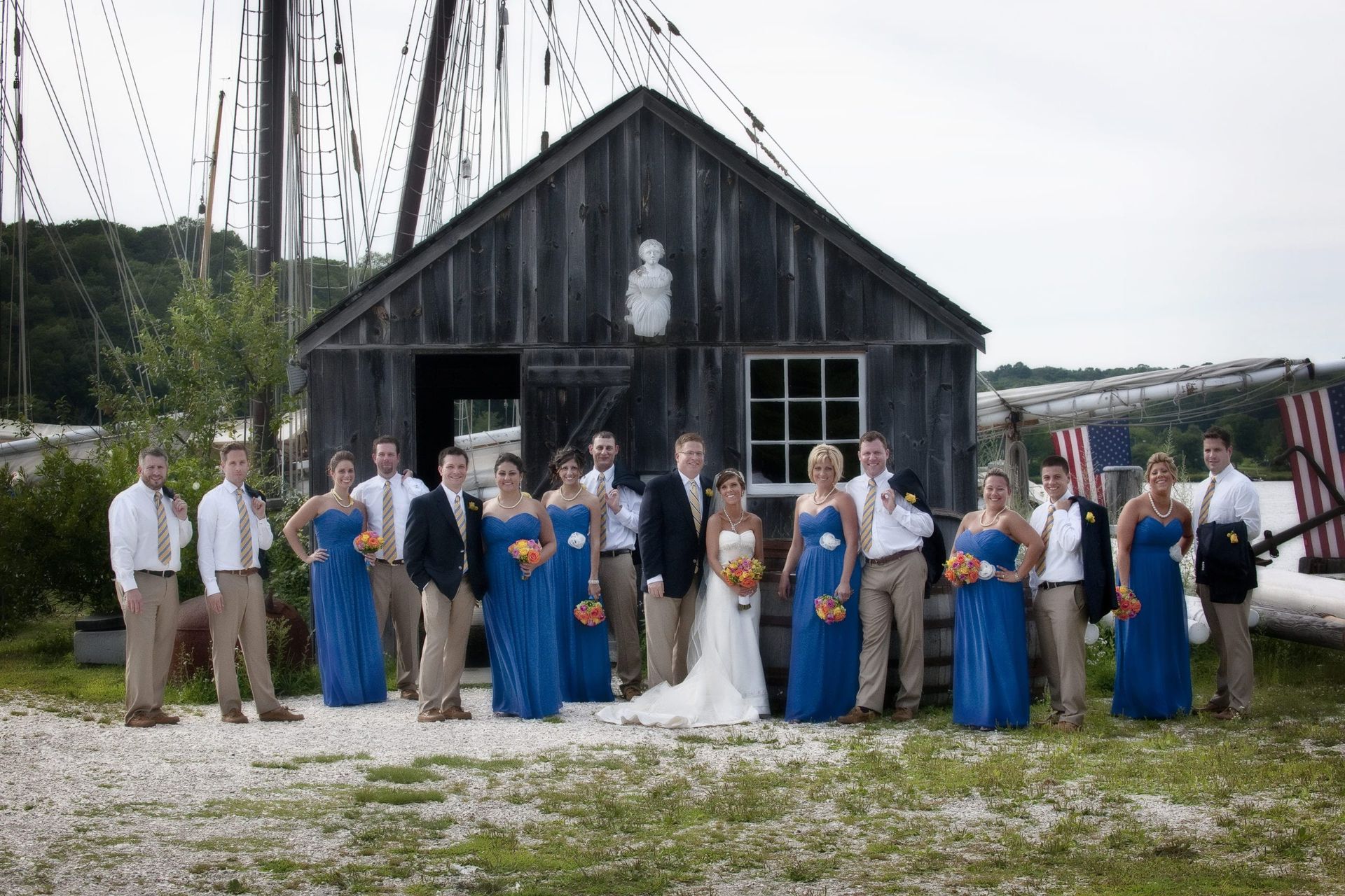 Wedding party in front of a weathered wooden building. Bride, groom, bridesmaids in blue, groomsmen in khaki.