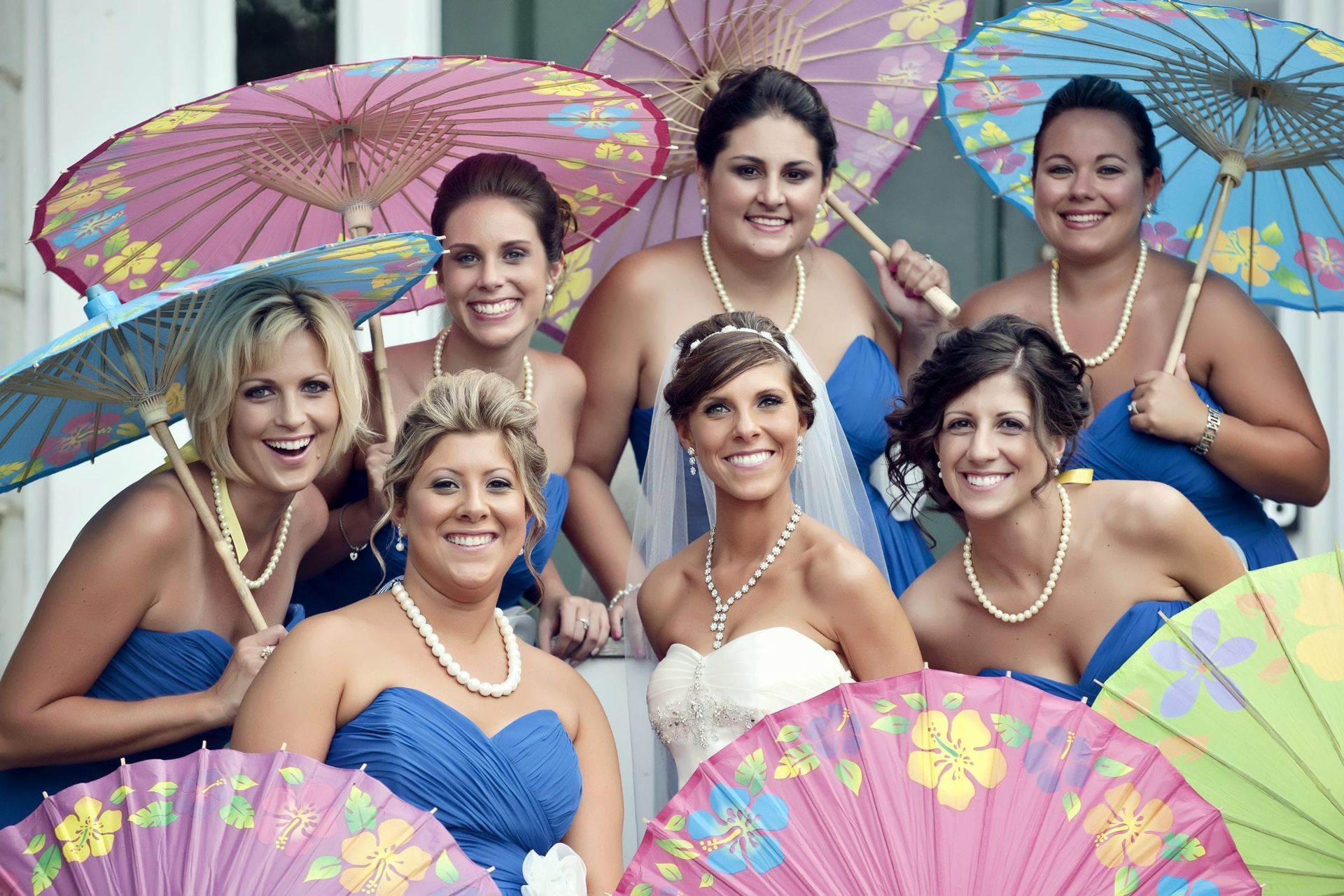 Bride and bridesmaids in blue dresses holding colorful parasols; all smiling outside.