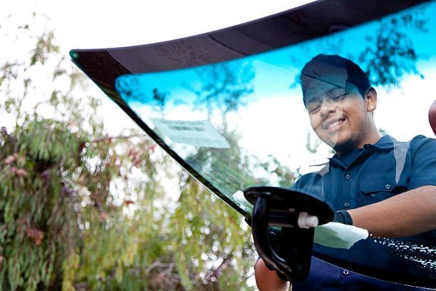 Person cleaning a car windshield with a blue microfiber cloth, smiling outdoors under a shaded canopy