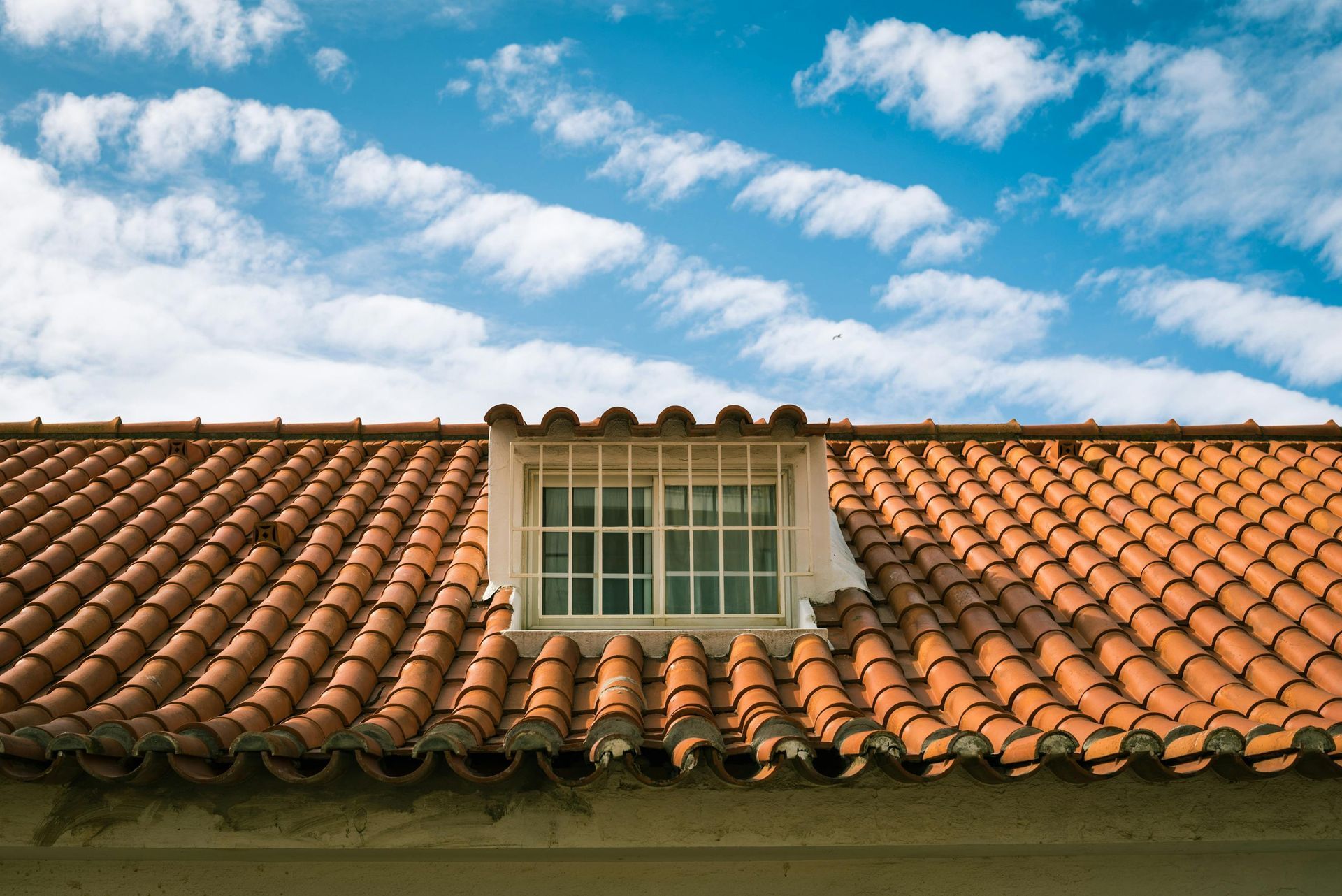 Red clay tile roof with a small white dormer window under a cloudy blue sky