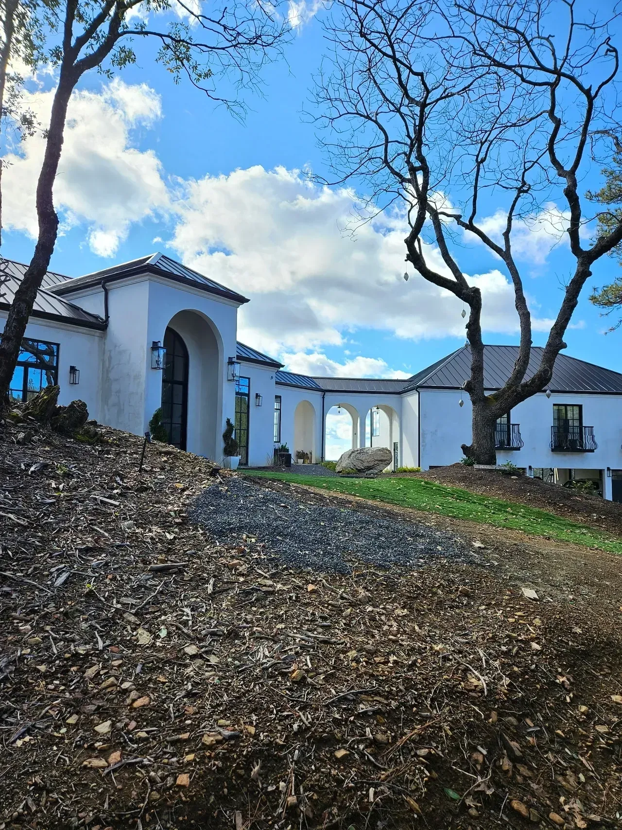 White stucco villa with blue trim on a hillside under a blue sky, framed by leafless trees and fallen leaves