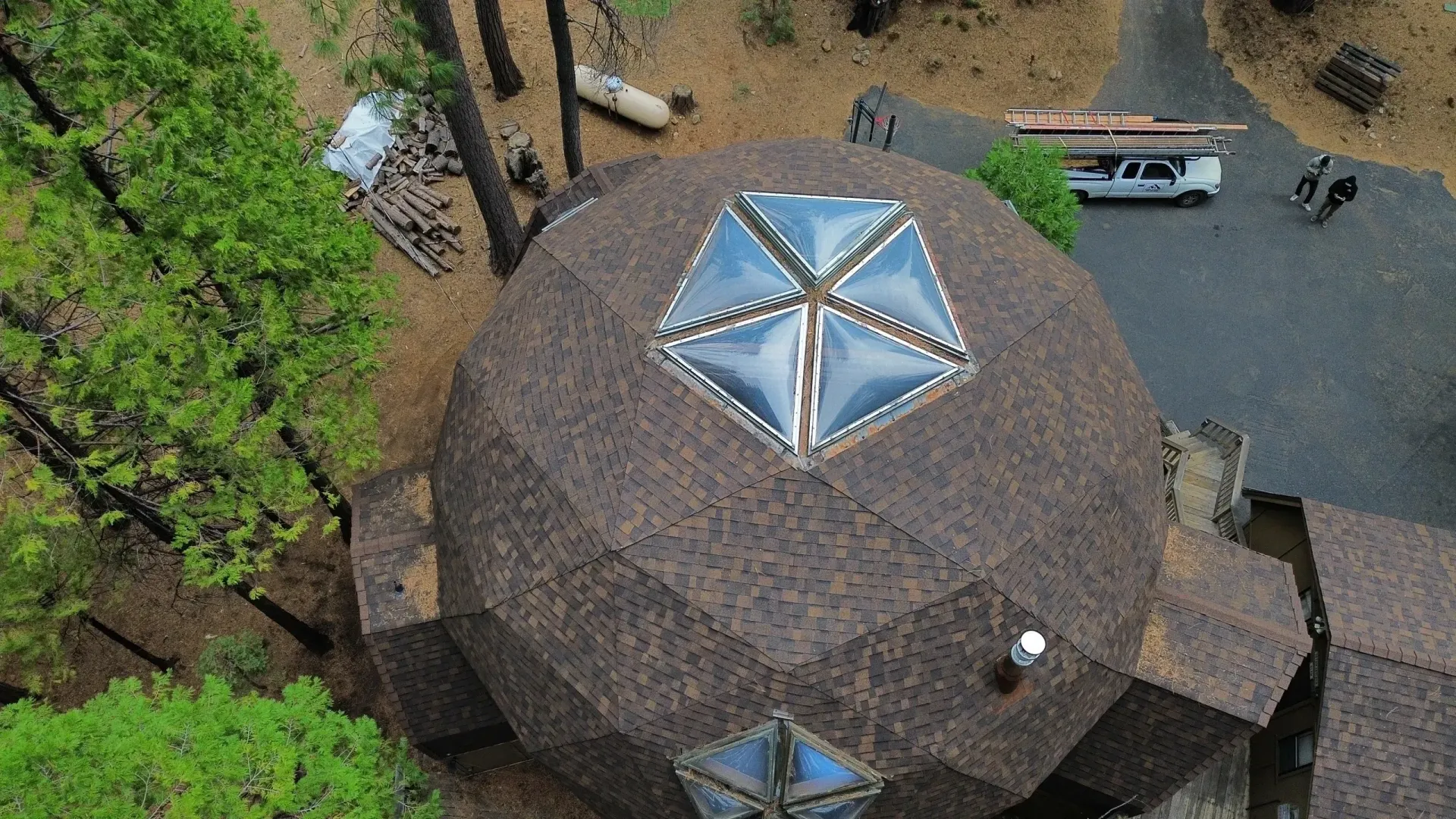 Aerial view of a round thatched-roof building with skylight windows surrounded by trees and a parking area.