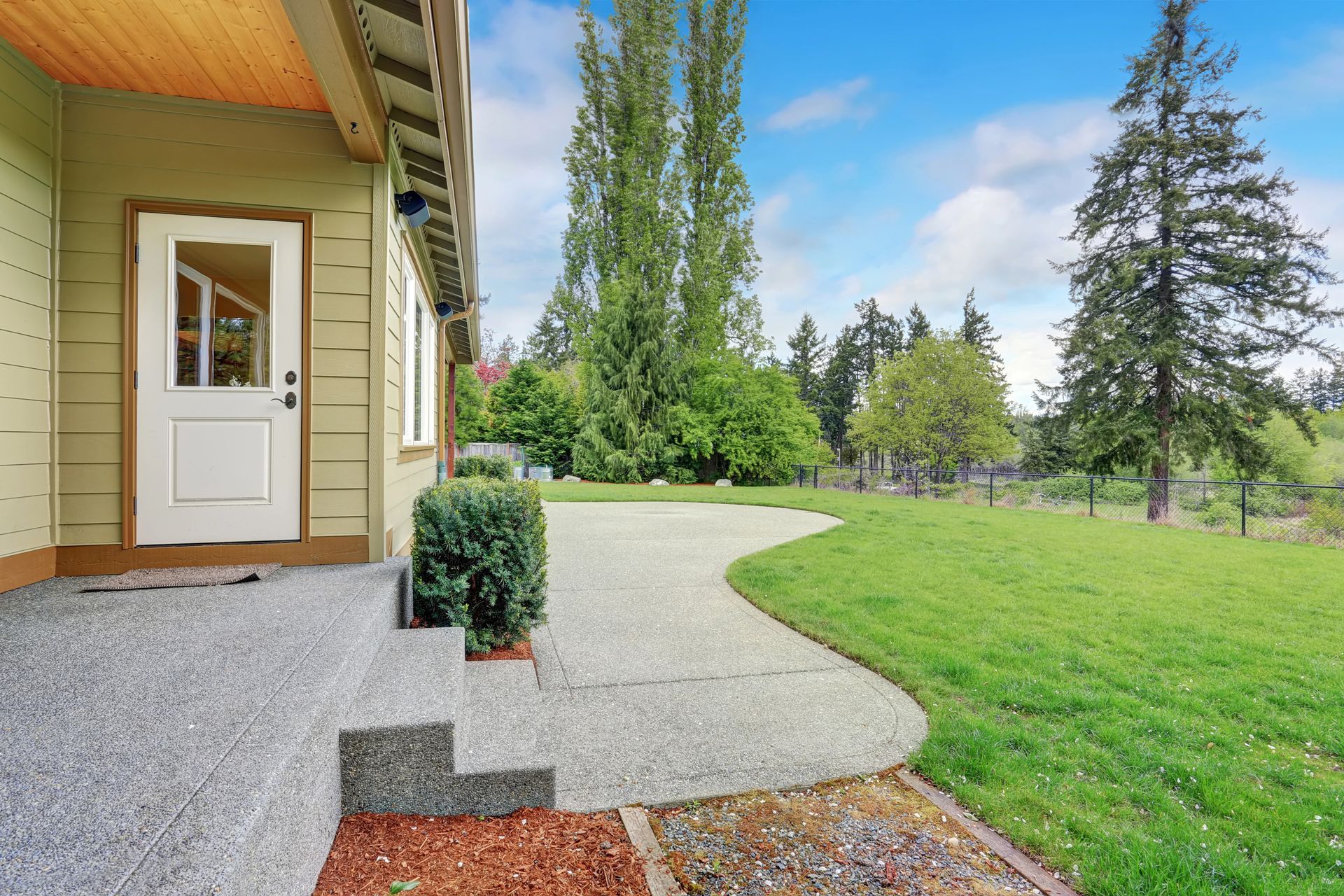 Exterior view of a house with a concrete patio, green lawn, and trees under a blue sky.