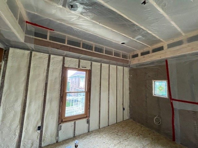 Interior of a room under construction, insulation between wooden beams and studs, a window, and a plastic sheet covering the ceiling.
