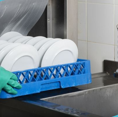 Person wearing green gloves places a blue rack of white plates into a commercial dishwasher.
