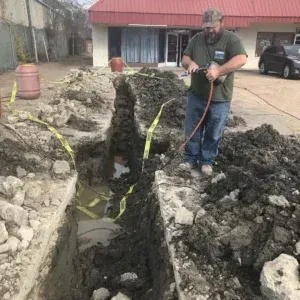 Man using a tool in a trench. Dirt and caution tape surround him. Building in the background.