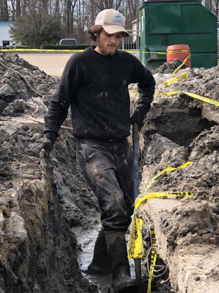 Man digging in a muddy trench, wearing a hat, long-sleeve shirt, and work boots. Yellow caution tape is visible.