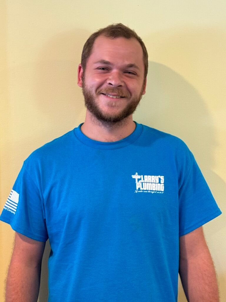 A man with a beard smiles, wearing a blue shirt with a logo. The background is a yellow wall.
