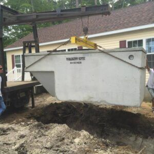 A concrete septic tank being lowered into a hole by a crane; men nearby, residential setting.