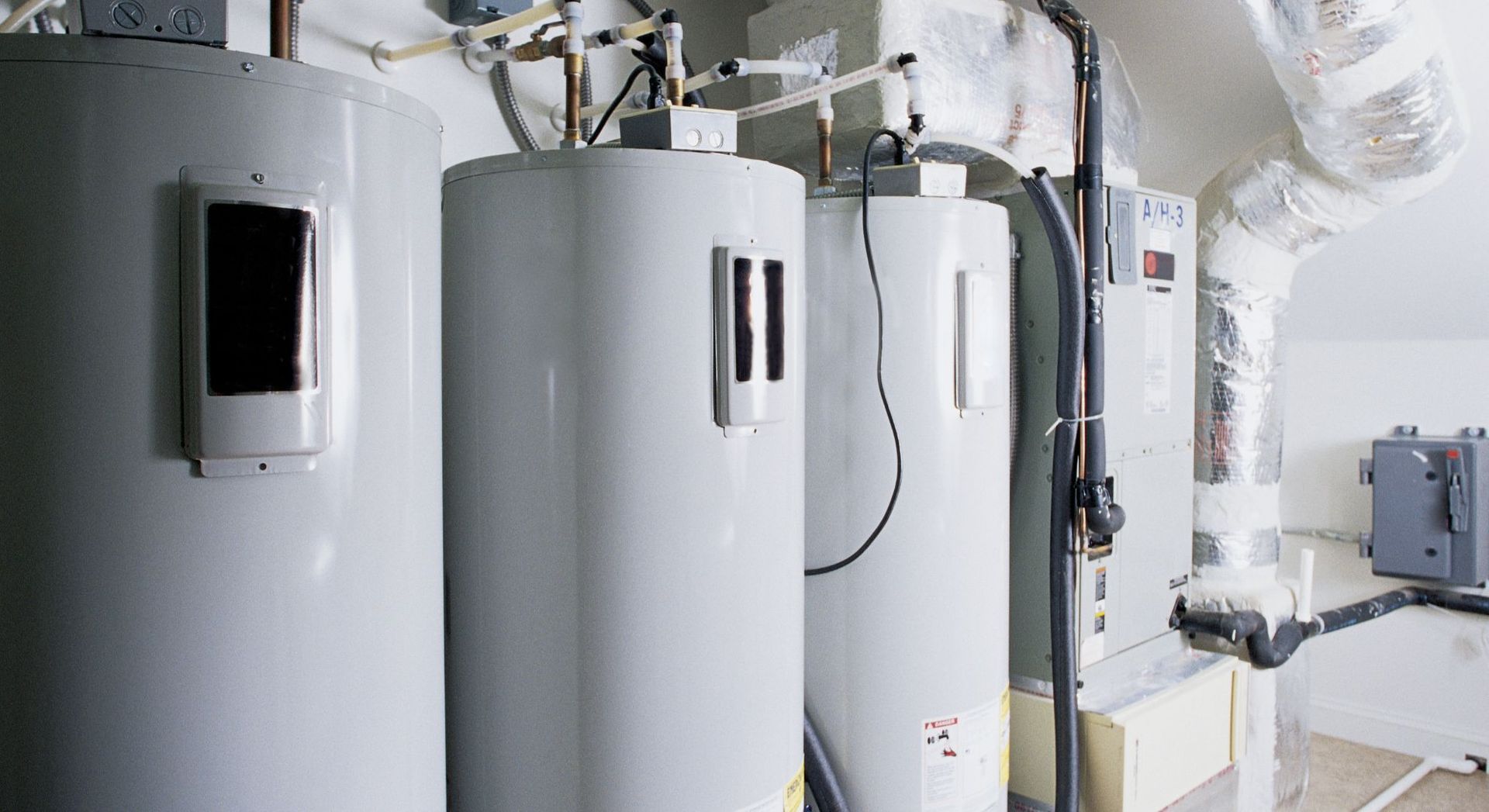 Three gray water heaters lined up in a room, with pipes and vents visible.