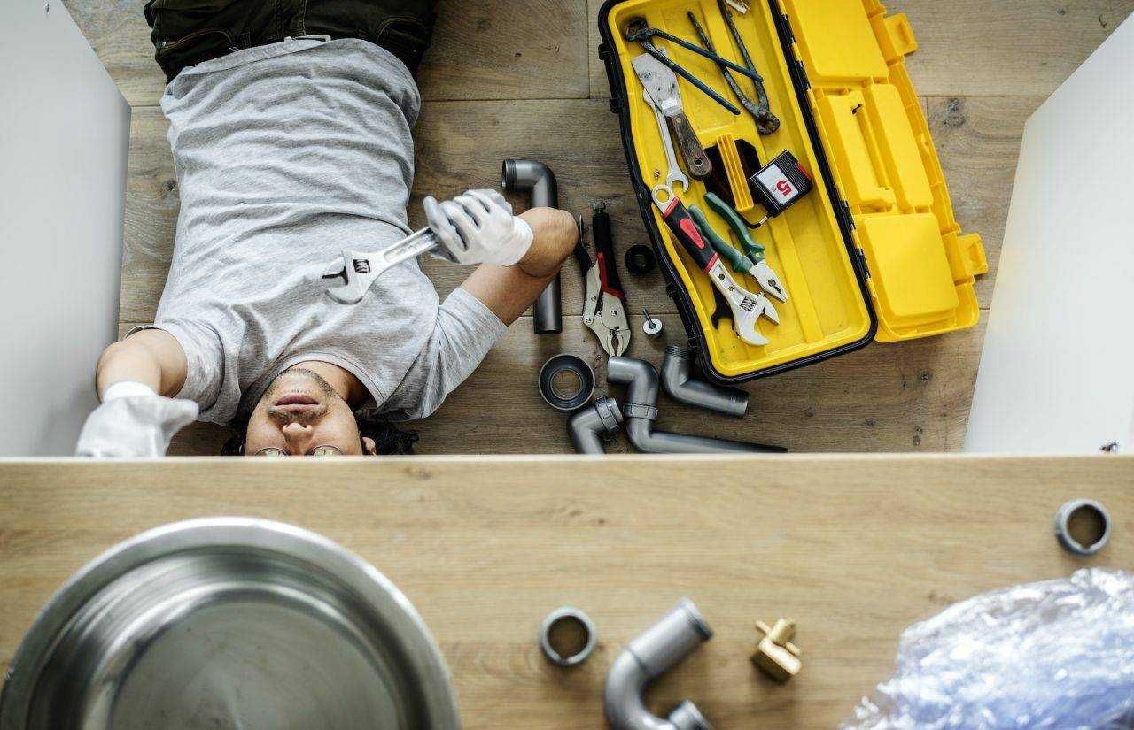 Plumber lying on the floor under a sink, working with tools from a yellow toolbox.