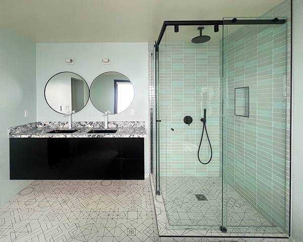 Modern bathroom with black vanity, round mirrors, glass shower with blue tile, and patterned floor.