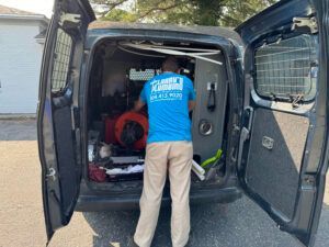 Plumber in blue shirt, beige pants, working inside a service van with the back doors open.