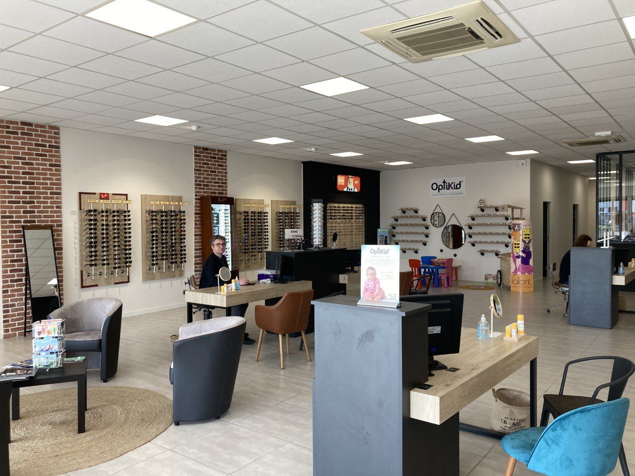 Intérieur d'un magasin d'optométrie avec des présentoirs à lunettes, des bureaux et une personne assise.
