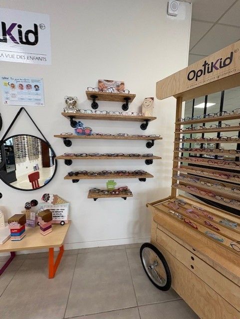 Intérieur d'un magasin de lunettes OptiKid avec des étagères présentant des lunettes et un chariot en bois.