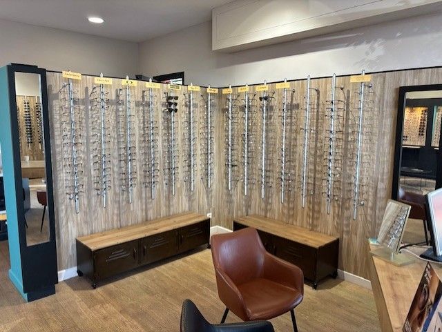 Des montures de lunettes exposées sur un mur en bois dans un magasin d'optique, avec des sièges et un miroir.