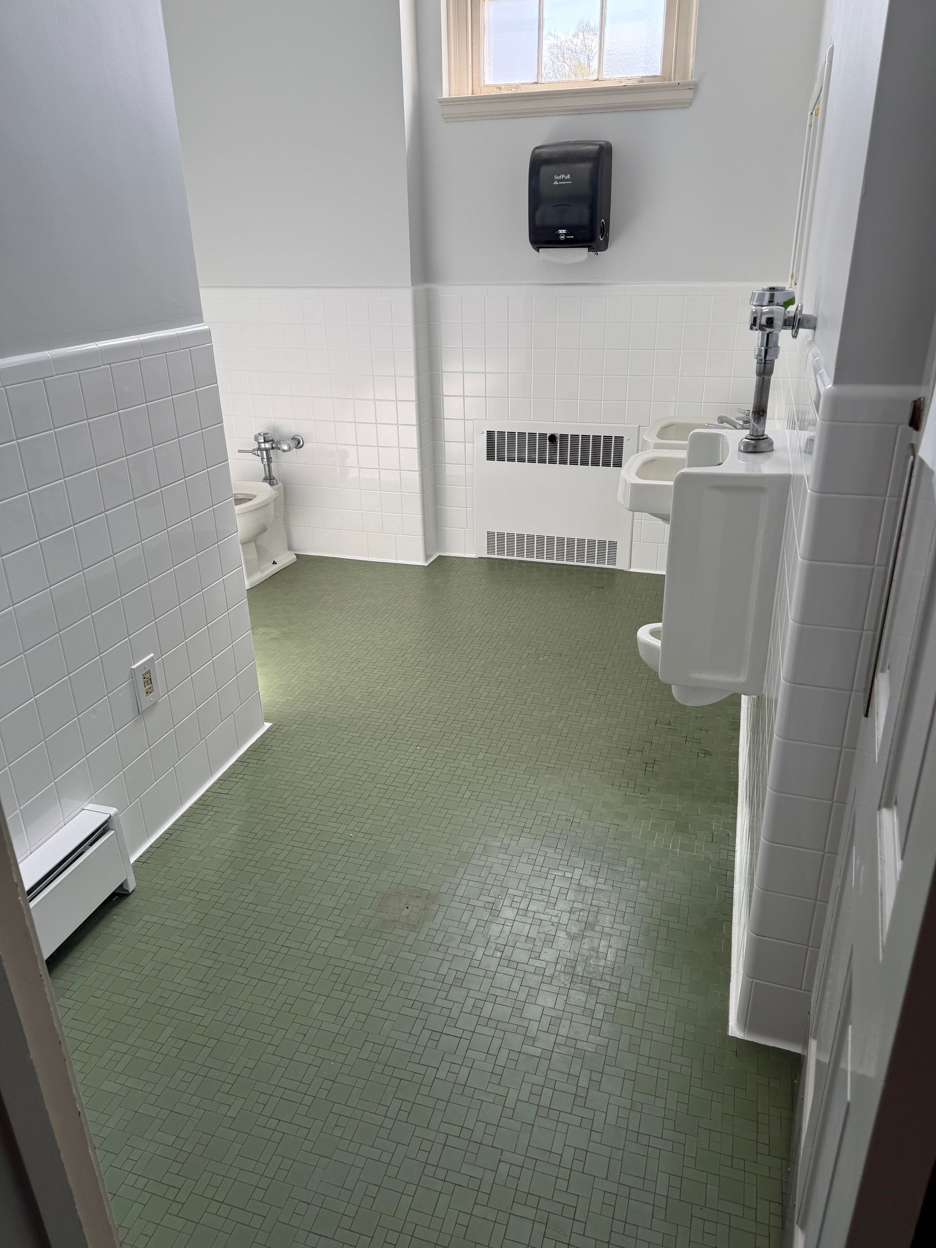 Small restroom with green floor, white tiled walls, urinal, sink, and soap dispenser near a window.