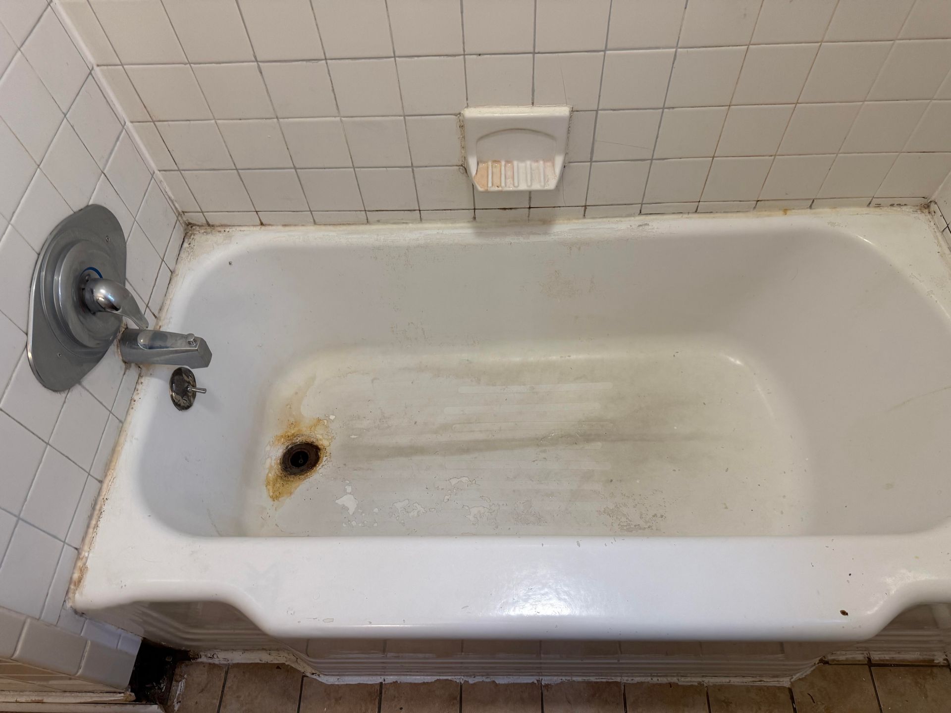 Dirty white bathtub with rust stains and a shower faucet in a tiled bathroom