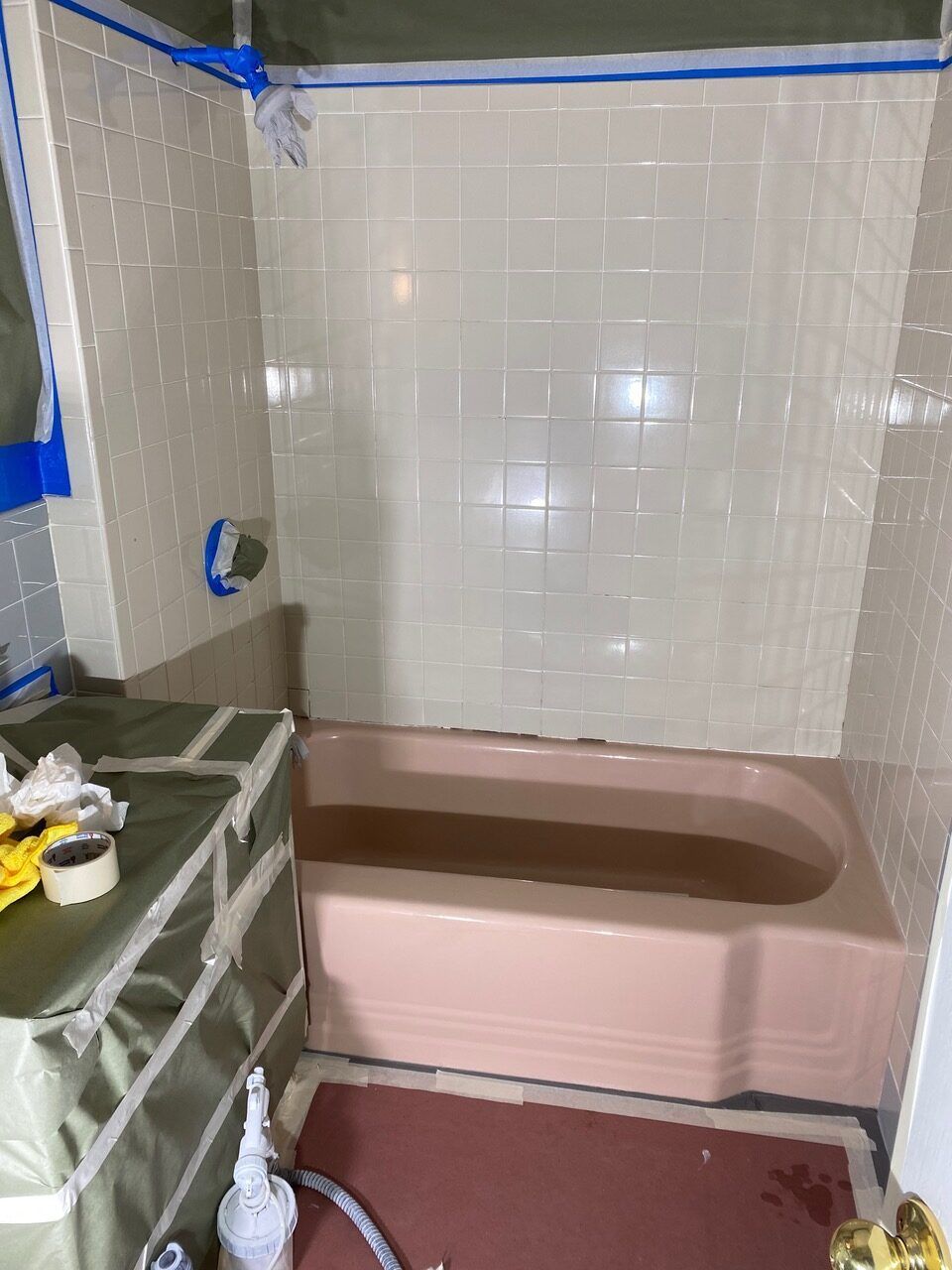 Pink bathtub and beige tiled walls in a bathroom being prepped for renovation with blue tape and protective coverings.