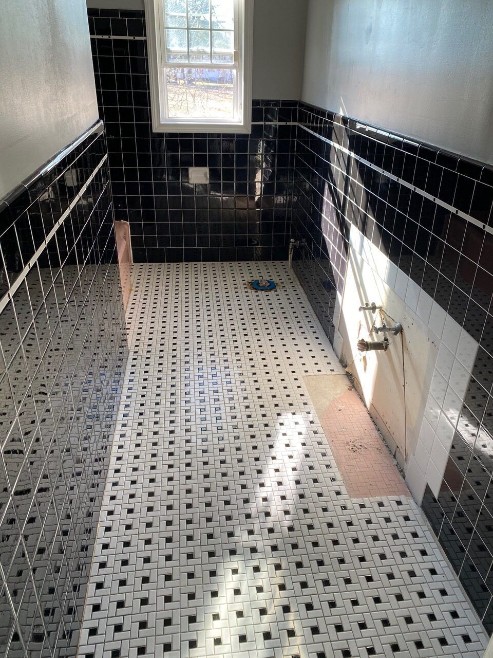 Bathroom under renovation: Black tiled walls, checkered floor. Partially tiled wall, exposed pipes, and a window.