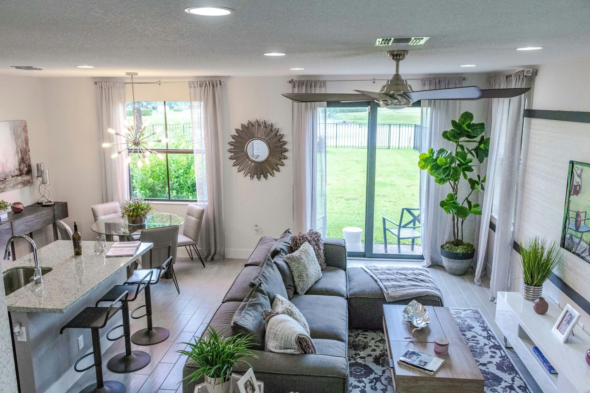 Open-concept living room with gray sectional, light-colored walls, large windows, and a view of a green lawn.