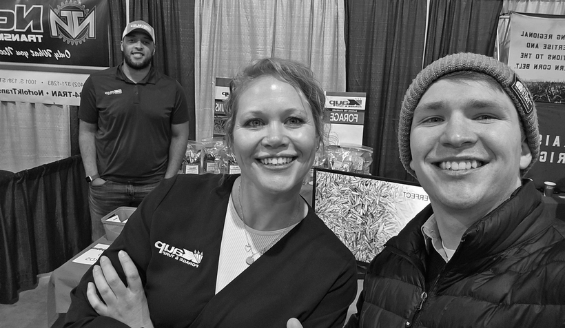 Three people smiling at a booth. Woman in scrubs and man in beanie stand front; man in cap stands behind.