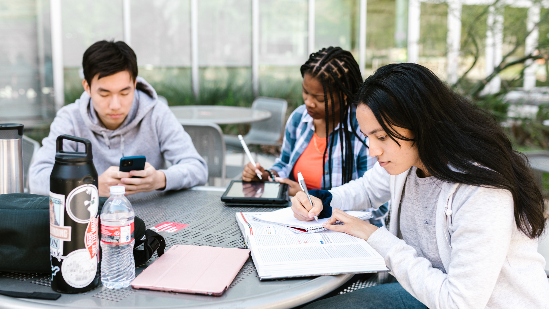 Three students studying outdoors at a table: one on phone, two writing, using tablet, water bottles present.