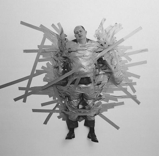 Man wrapped in foil and tape, with wooden sticks radiating outward. Black and white.