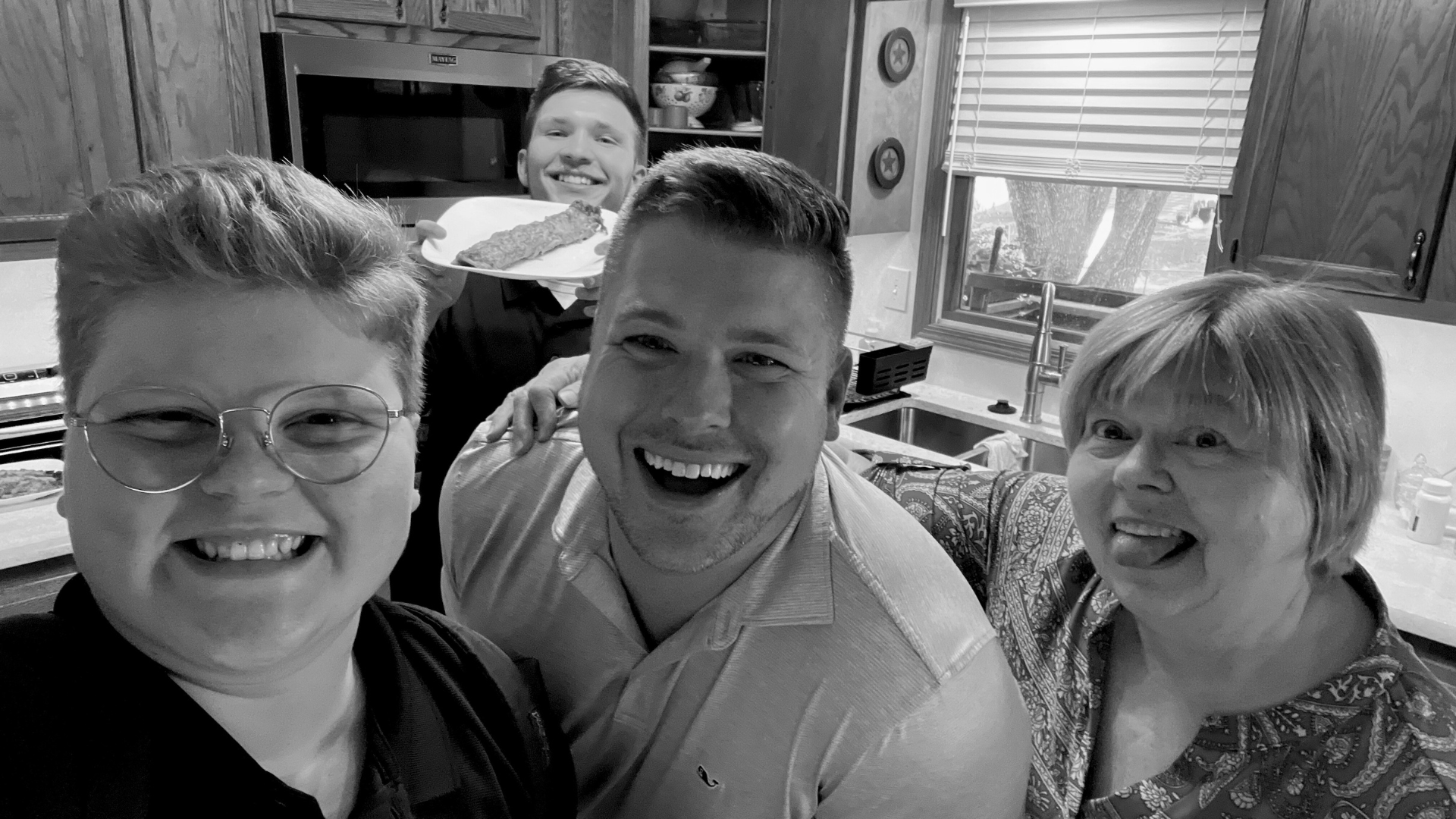 Four smiling people in a kitchen. One holds a tray. Black and white.