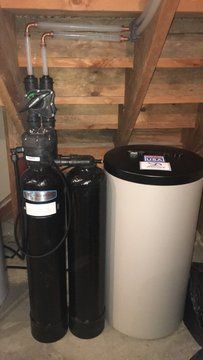 A water softener is sitting under a wooden staircase in a basement.